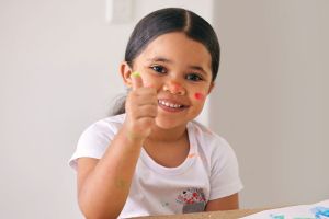Smiling young girl holding thumbs up while fingerpainting