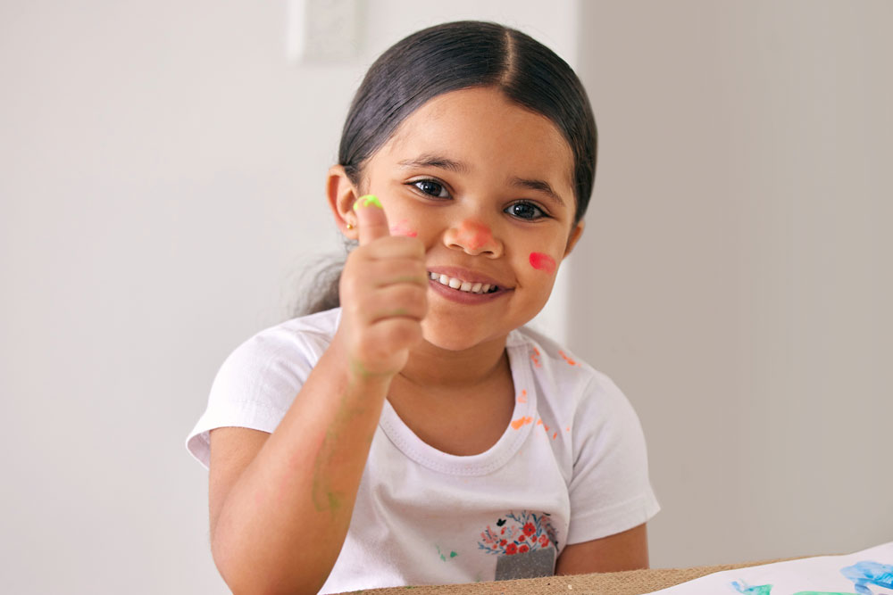 Smiling young girl holding thumbs up while fingerpainting