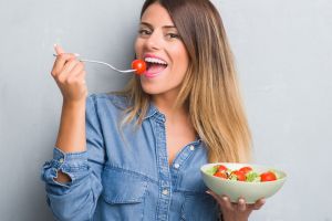 Smiling young woman eating salad on gray background